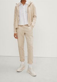 Beige hoodie met rits en trekkoord, witte blouse met kraag, beige cargobroek met zijzak, en cremewitte sneakers met een gestructureerde zool.
