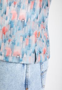 Camisa de manga corta con un patrón de horizonte urbano en azul y rosa, con palmeras. Fabricada en una mezcla de algodón con una etiqueta lateral. Se ven pantalones cortos de mezclilla.