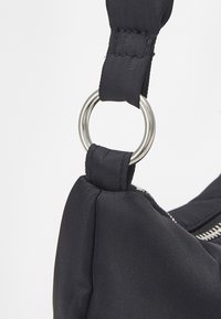 Czarna torba z nylonu o teksturowej powierzchni, z metalowym srebrnym pierścieniem na pasku i detalem w postaci zamka. Czyste linie i praktyczny design.