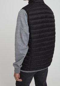 Schwarze Steppweste mit gequilten Abschnitten, hohem Kragen und glatter Oberfläche, getragen über einem grauen Langarmshirt. Seitenansicht dargestellt.