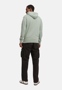 Sudadera con capucha verde menta, con puños y dobladillo acanalados, combinada con pantalones cargo negros con bolsillos laterales, y sandalias ligeras.