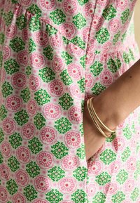 Puuvillane kleit, millel on roosa ja rohelise lillemuster, koos kogutud detailiga vööl ja kahe küljetaskuga. Kuldsed käevõrud randmel.