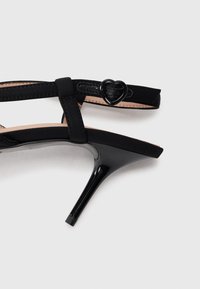 Sandală neagră cu toc înalt, cu un finisaj patent elegant, având o curea subțire și o cataramă în formă de inimă. Branțul texturat completează designul.