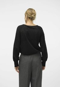 Sort struktureret strikbluse med lange puffærmer, parret med grå højtaljede bukser, set fra ryggen. Simpelt design, afslappet pasform.
