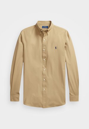Camisa de manga longa bege clara, com botões, feita de algodão suave. Apresenta um colarinho pontudo e um pequeno logótipo de cavalo azul bordado no peito.