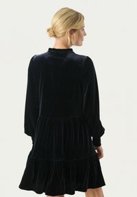 Robe en velours noir avec manches longues, ourlet volé et col haut. La texture est douce, avec un léger éclat et des détails froncés.