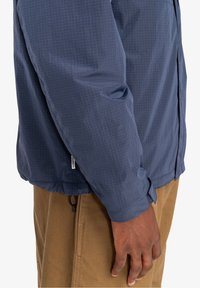 Granatowa lekka kurtka z wzorem w kratę i regulowanymi mankietami, w zestawieniu z beżowymi spodniami. Kurtka posiada sznurek do regulacji na dole.