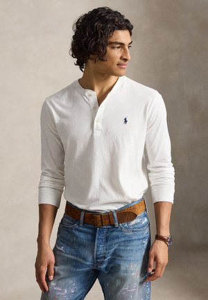 Homme aux cheveux bouclés portant un t-shirt henley blanc à manches longues, un jean bleu éclaboussé de peinture, une ceinture marron et une montre-bracelet marron regardant vers la droite.