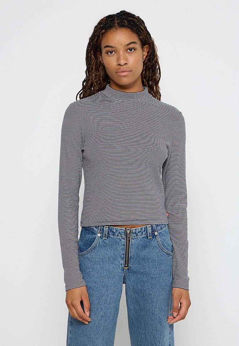 Langärmliges, hochgeschlossenes Oberteil in grau und weißen horizontalen Streifen, aus weichem Stoff. Kombiniert mit hochgeschnittenen blauen Jeans.