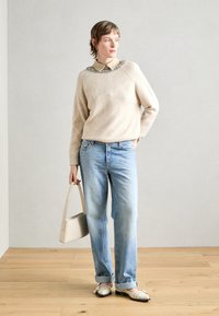 Pull en maille beige clair avec col décoratif, associé à un jean droit bleu clair et des chaussures blanches. Tient un petit sac à main beige.