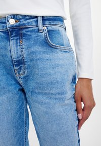 Person som bär ljusblå denimjeans och en vit långärmad topp, med höger hand vilande vid sidan.