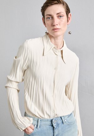 Crème plooiblouse met een puntige kraag en knoopsluiting, gecombineerd met lichtblauwe denim jeans. Zachte stof en lange mouwen met opgerolde manchetten.