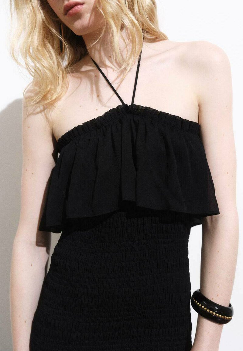 Svart strapless klänning med volangdetaljer, smockat utseende och en figurnära silhuett. Accessoarer med ett svart armband.