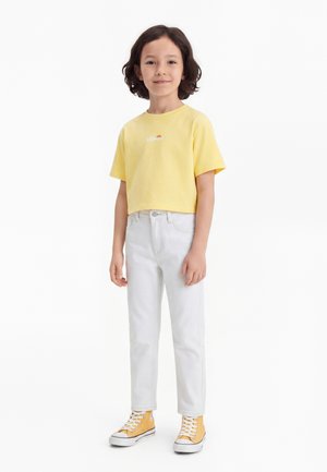 Bambino con capelli scuri fino alle spalle che indossa una maglietta gialla corta, jeans bianchi e scarpe da ginnastica alte gialle, in piedi su sfondo bianco.