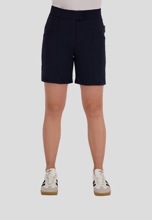 Persoon in zwarte shorts en witte sneakers met zwarte strepen, staand tegen een effen grijze achtergrond.