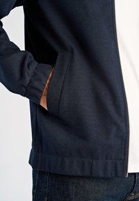 Granatowa kurtka na zamek wykonana z gładkiej tkaniny, z bocznymi kieszeniami, elastycznymi mankietami i szczegółami przeszyć, zestawiona z ciemnymi jeansami z denimu.