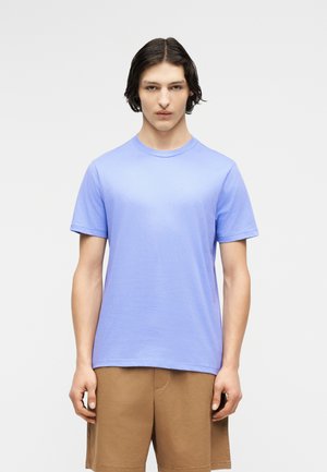 Młoda osoba z ciemnymi włosami do ramion, ubrana w jasnoniebieską krótką koszulkę i brązowe szorty na jednolitym tle.