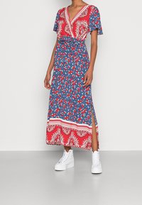 Maxiklänning i blått, rött och vitt paisley-mönster, med en omslagdesign, korta ärmar och sidoklyfta, som paras ihop med vita chunky sneakers.