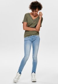 Camiseta verde de cuello en V combinada con jeans ajustados azul claro y zapatillas blancas, mostrando un ajuste casual y una textura de tela suave.
