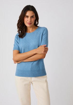 Mujer con cabello oscuro que lleva un suéter azul claro de manga corta y pantalones color crema, de pie con los brazos cruzados frente a un fondo liso.