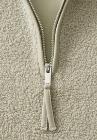 Beige mit Reißverschluss, ausgestattet mit einem silbernen Metallreißverschluss und weichem, strukturiertem Stoff, mit einem glatten Ziehband. Im Hintergrund ist ein flauschiges Material zu sehen.