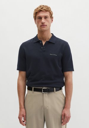 Blonder Mann trägt ein marineblaues kurzärmeliges Polo-Shirt mit dem Schriftzug „Marc O’Polo“, beige Hose und schwarzen Gürtel, steht vor einem einfarbigen hellen Hintergrund.