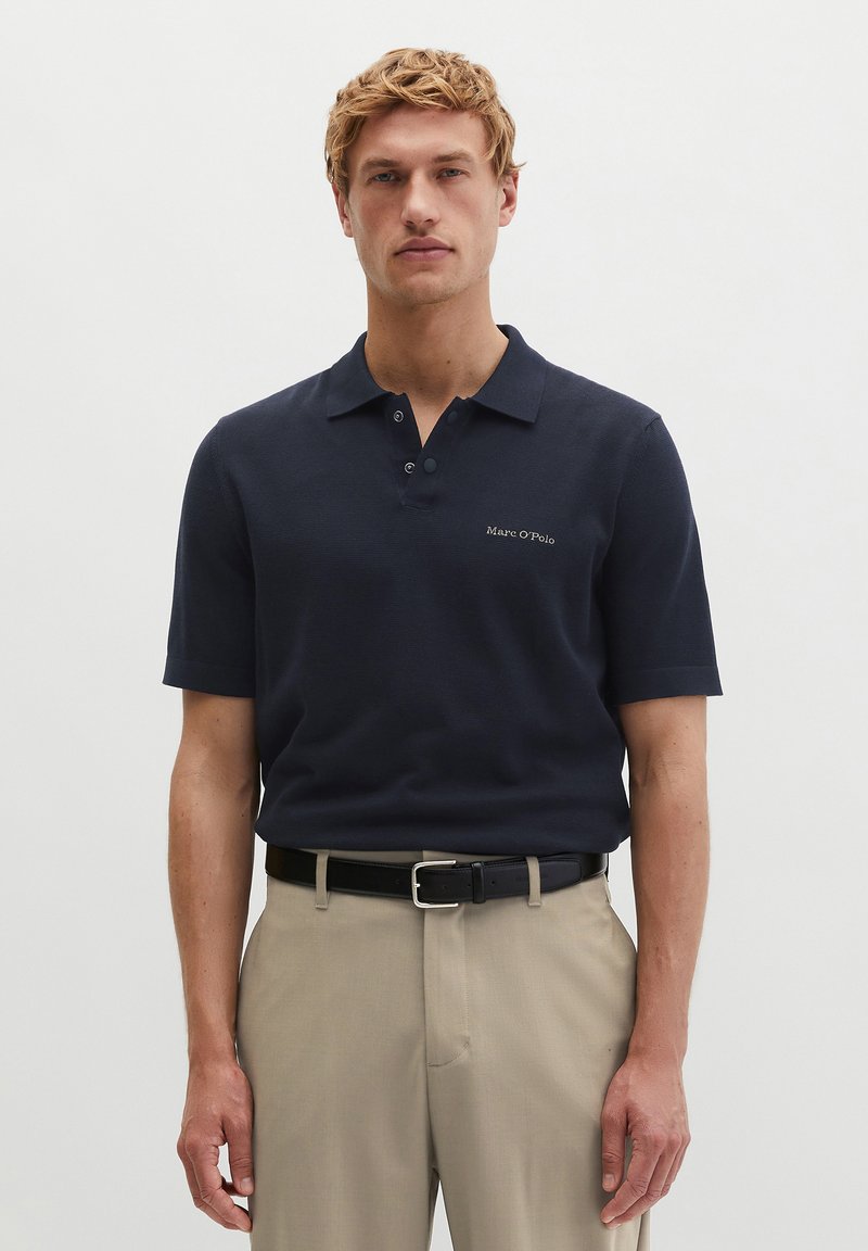 Uomo biondo che indossa una polo blu navy a maniche corte con la scritta "Marc O’Polo", pantaloni beige e cintura nera, in piedi davanti a uno sfondo chiaro uniforme.