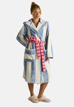 Frau, die einen blau-weiß gestreiften Kapuzenmantel mit einem pink gestreiften Gürtel und beigen Hausschuhen mit Pelzfutter trägt, steht vor einem schlichten Hintergrund.