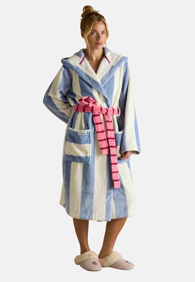 Naine kannab sinise ja valge triibulist kapuutsi ning roosa triiburihma, samuti beeže labakuid, mille sees on karusnahk, seismas ühtlase tausta taustal.