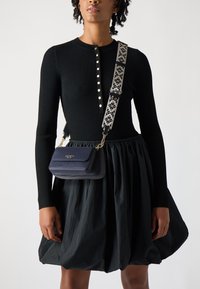 Sac en cuir marine avec une finition texturée, quincaillerie dorée, et bandoulière à motif floral. Porté avec un pull noir à côtes et une jupe noire.