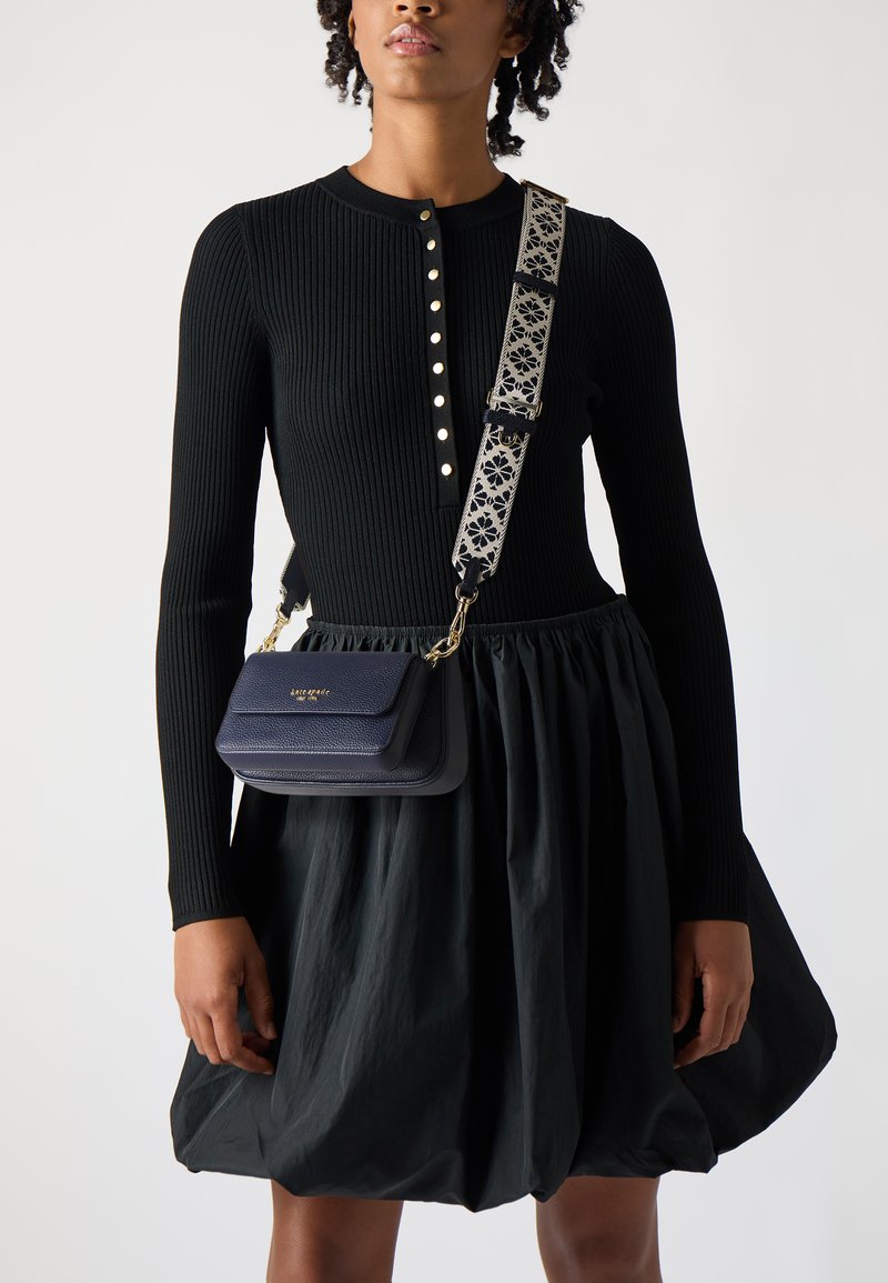 Sac en cuir marine avec une finition texturée, quincaillerie dorée, et bandoulière à motif floral. Porté avec un pull noir à côtes et une jupe noire.