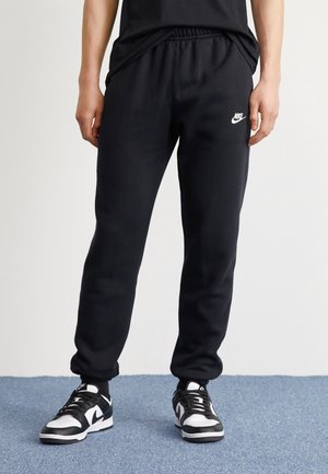 Person trägt schwarze Nike Jogginghose mit elastischen Bündchen und schwarz-weiße Nike Sneakers, steht auf blauem Teppich.