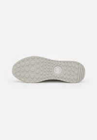 Semelle de chaussure gris clair avec des motifs circulaires texturés et un logo au centre, présentant une finition lisse et un talon strié pour une meilleure adhérence.