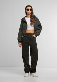 Felpa corta grigio scuro con zip frontale, abbinata a pantaloni neri larghi e sneakers bianche. Gli occhiali da sole completano l'outfit.