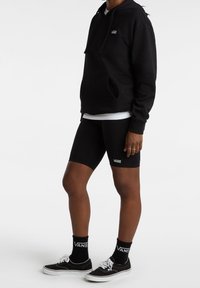 Czarna bluza z kapturem z przednią kieszenią, zestawiona z czarnymi szortami rowerowymi i czarnymi sneakersami. Logo Vans widoczne na odzieży i skarpetach.