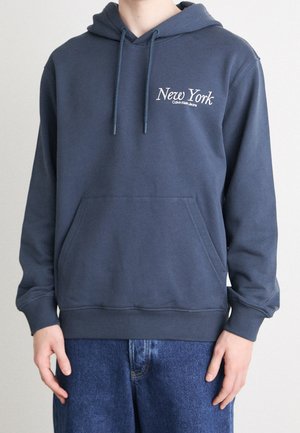 Person trägt einen marineblauen Hoodie mit dem Schriftzug "New York Calvin Klein Jeans", kombiniert mit blauen Denim-Jeans, dargestellt vom Hals bis zu den Hüften.