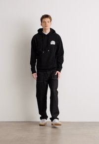 Sudadera con capucha negra y un gráfico en la parte frontal, combinada con pantalones de mezclilla oscuros y zapatillas blancas, de pie contra una pared clara.