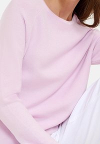 Kvinna som bär en ljusrosa ribbad långärmad tröja och vita byxor, med hår synligt på ena sidan.