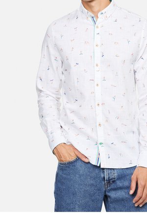 Man wearing white button-up shirt with small colorful swimmer illustrations and blue jeans, hand in pocket.