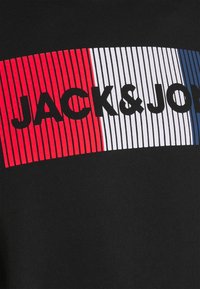 Camiseta negra con el texto "JACK&JONES" en letras grandes en rayas negras, blancas y rojas en la parte frontal. Material de algodón con una textura suave.