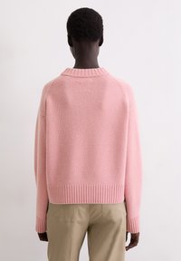 Eine Person mit dunkler Haut und kurzen, lockigen Haaren trägt einen hellrosa Strickpullover und beige Hosen und steht mit dem Rücken zu einem einfarbigen Hintergrund.