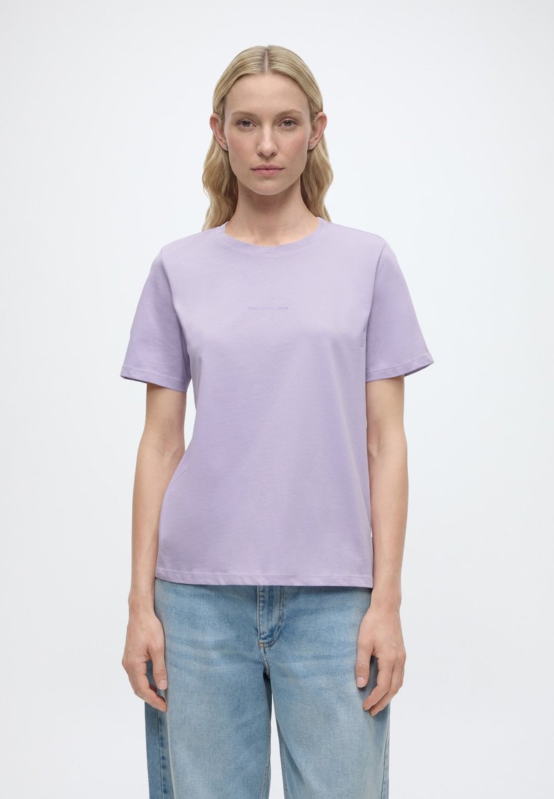 Ξανθιά γυναίκα με ίσια μαλλιά φοράει απλή λιλά κοντομάνικη μπλούζα και ανοιχτό μπλε τζιν, στέκεται μπροστά σε λευκό φόντο.