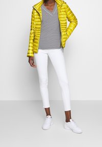 Giacca a vento gialla con finitura lucida, maglietta a strisce con scollo a V, pantaloni bianchi aderenti e sneakers bianche. Design semplice e vestibilità moderna.