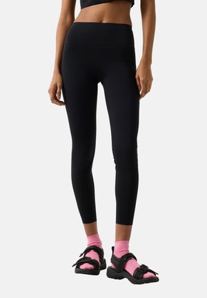 Kvinde iført sorte leggings med høj talje, lyserøde sokker og sorte sporty sandaler, stående foran en hvid baggrund.