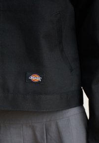 Veste en toile noire au design cropped, dotée d'une poche latérale et d'un patch logo Dickies bien visible. Texture lisse avec des coutures soignées.