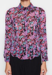 Blusa floral transparente con fondo negro y flores rosas, moradas y azules. Diseño con cuello, cierre de botones y mangas largas.