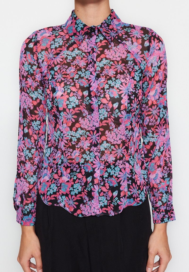 Blusa floral transparente con fondo negro y flores rosas, moradas y azules. Diseño con cuello, cierre de botones y mangas largas.