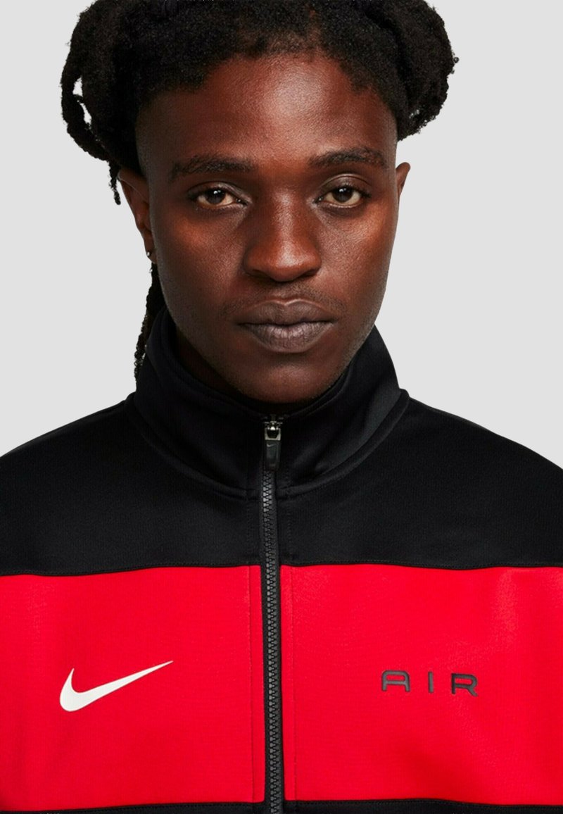 Adulto con piel oscura y cabello trenzado que lleva una chaqueta Nike negra y roja con cremallera, con el logo blanco de swoosh y "AIR".