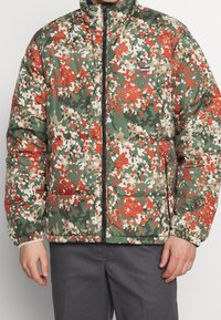 Homme portant une doudoune zippée avec un motif camouflage rouge, vert et beige, pantalon noir, les mains détendues le long du corps.