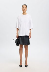 Hvid oversized t-shirt, sort lædernederdel, sorte spidse hæle, holder en lille sort håndtaske, korte ærmer, enkelt design, glat tekstur.